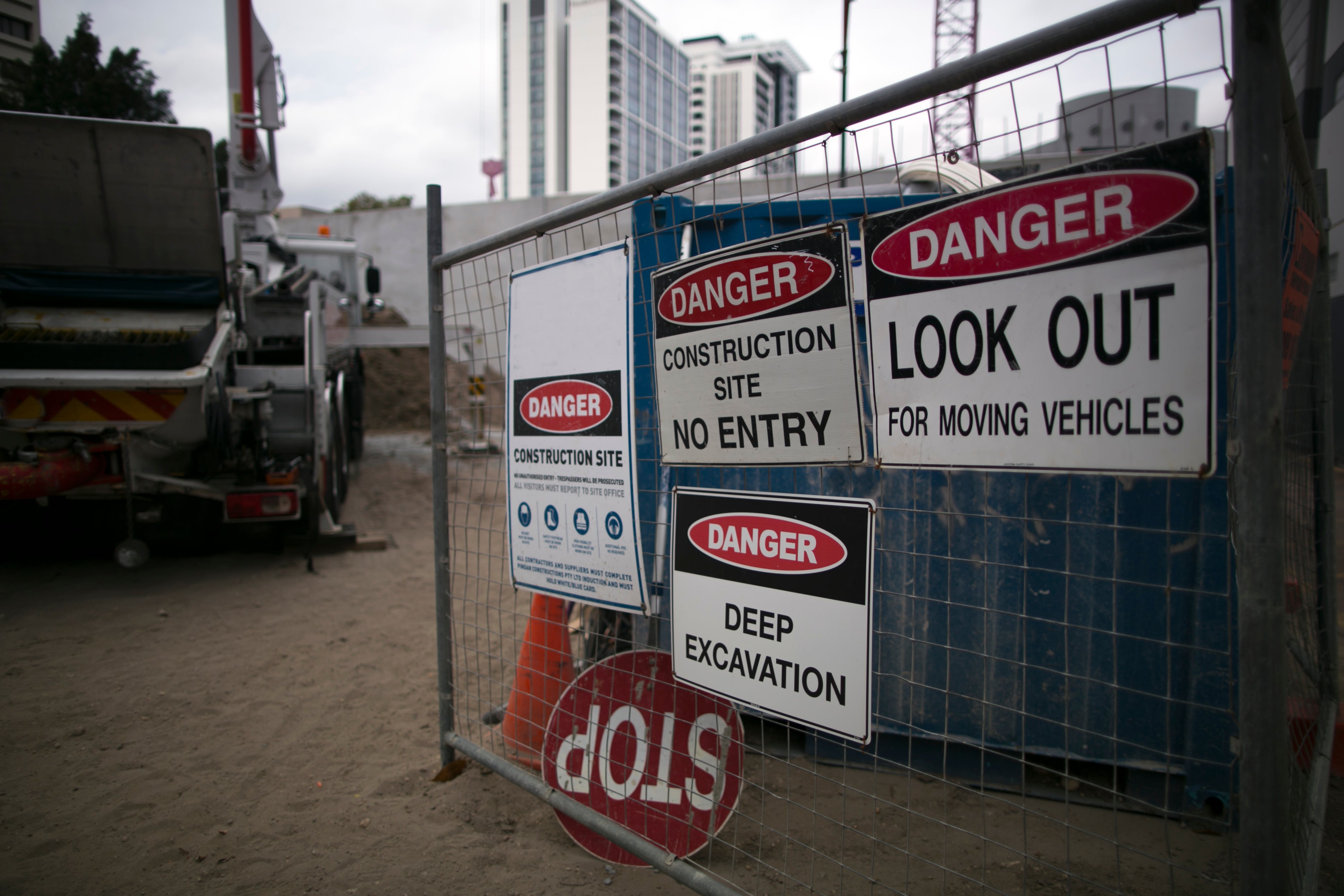 Construction site entrance with safety signs displayed on a metal fence, including 'Danger - Construction Site No Entry,' 'Look Out for Moving Vehicles,' and 'Deep Excavation,' alongside a visible stop sign and construction machinery in the background